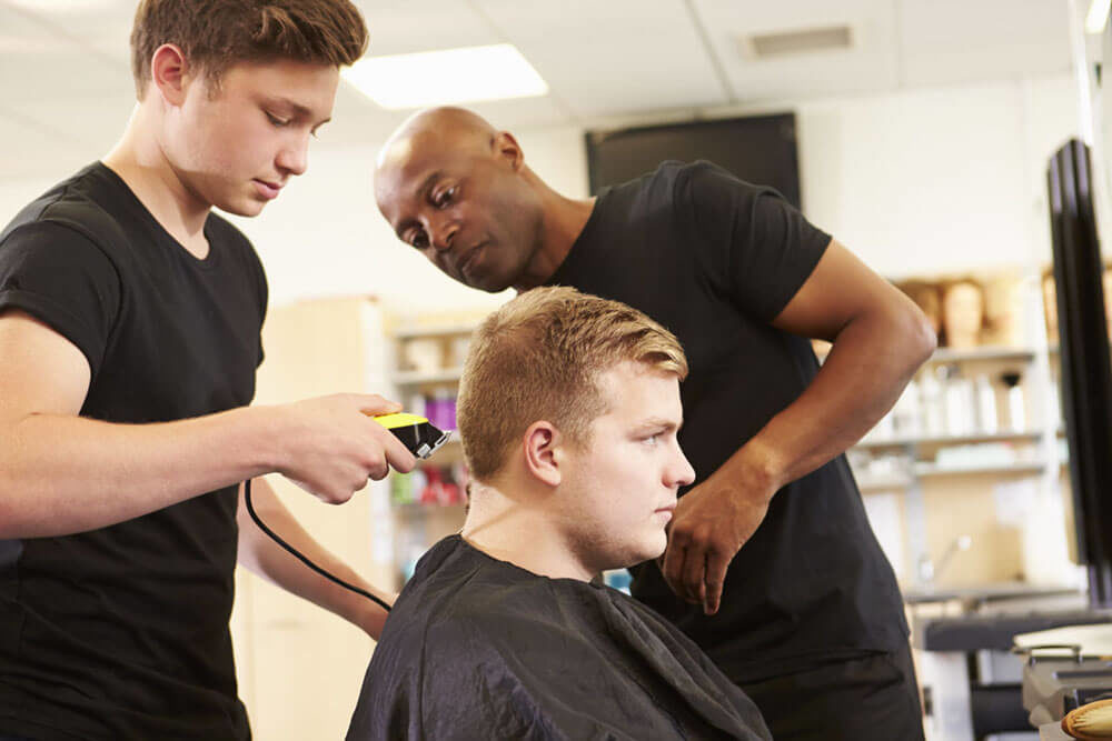 Trois hommes dans un salon de coiffure, dont un qui se fait raser les cheveux par un apprenti supervisé par un coiffeur qui semble plus expérimenté.